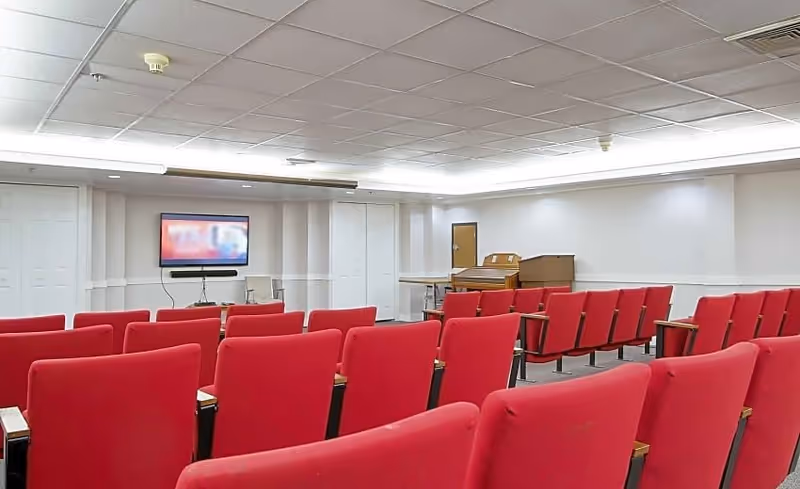 Rows of red upholstered theater-style chairs facing a wall-mounted TV and a piano in a light-colored activity/meeting room.