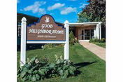 Outdoor view of the Good Neighbor Home main entrance sign with a building entrance and pathway in the background under a blue sky with some clouds.