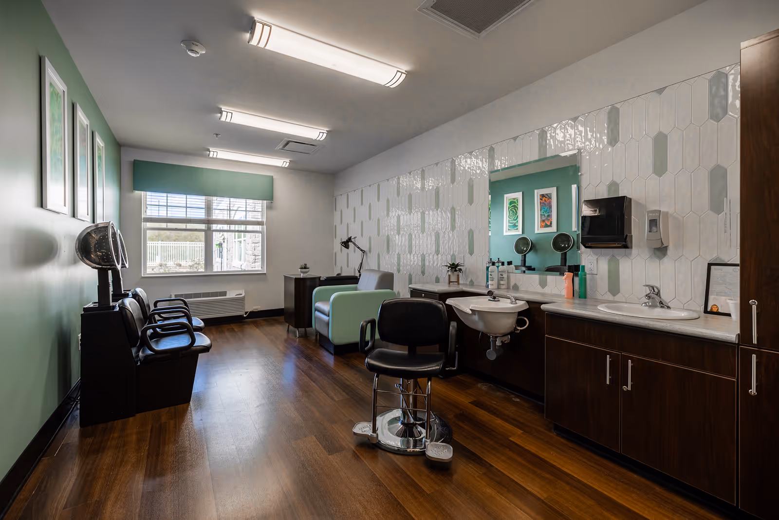 Interior of a salon room with wooden flooring, a green accent wall with framed artwork, three black salon chairs with hair dryers, a mint green armchair, a large mirror with white and green tile backsplash, a sink, and cabinets.