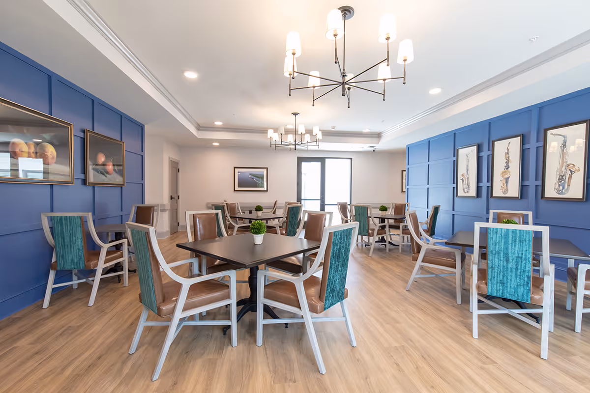 A bright dining room with multiple square tables surrounded by chairs with teal and brown upholstery. The room features blue paneled walls adorned with framed artwork, light wood flooring, and modern chandeliers hanging from the ceiling. A door with glass panels is visible at the far end of the room.