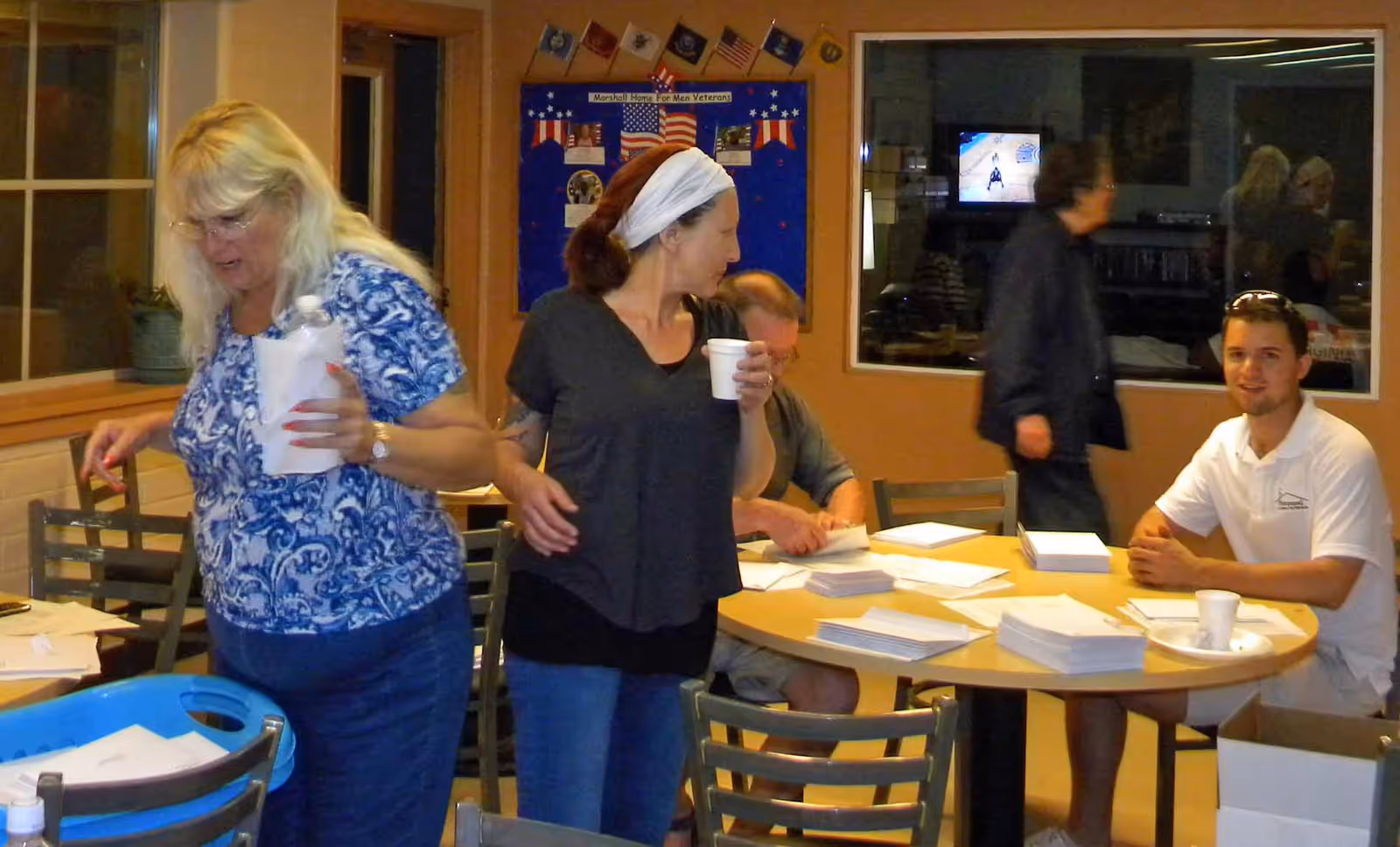 Several people gathered around a round table in a communal dining/activity room sorting stacks of paper and drinking from cups.