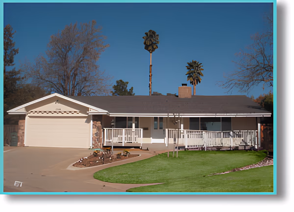 Single-story residential building with a front porch, white railing, and a two-car garage. The house is surrounded by a lawn and some trees under a clear blue sky.