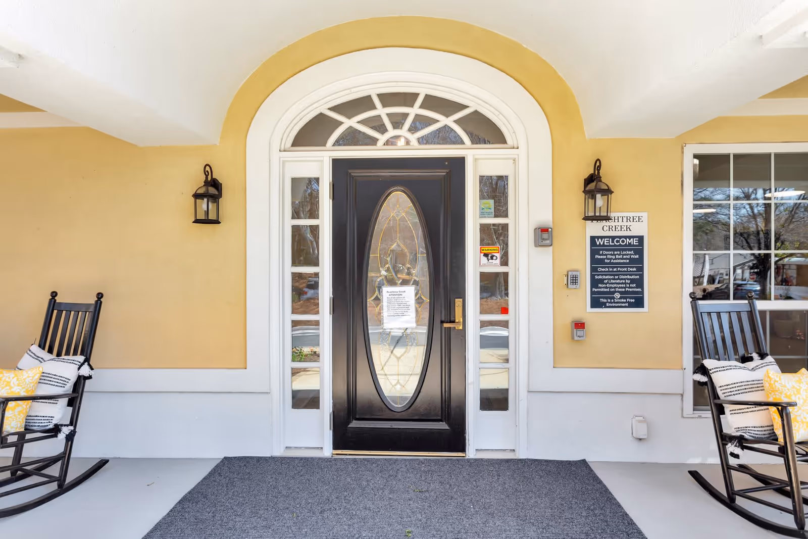 Entrance door of Peachtree Creek Memory Care facility with a black door featuring an oval glass panel, flanked by sidelights and topped with a fanlight window. Two black rocking chairs with yellow and white cushions are placed on either side of the door. A welcome sign with facility rules is mounted on the right wall next to the door.