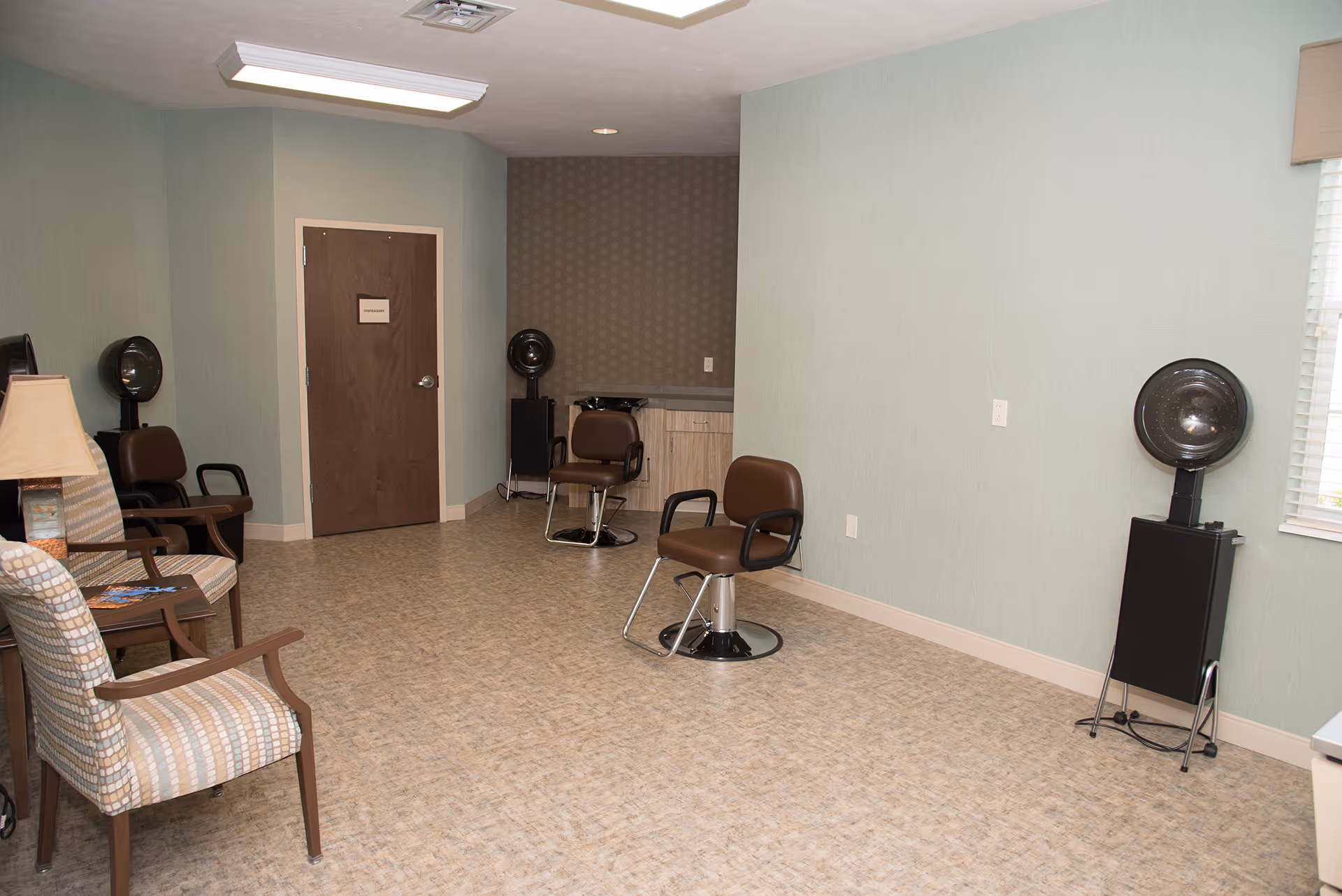 A small salon-style room with brown styling chairs and hooded hair dryers in a senior living facility.