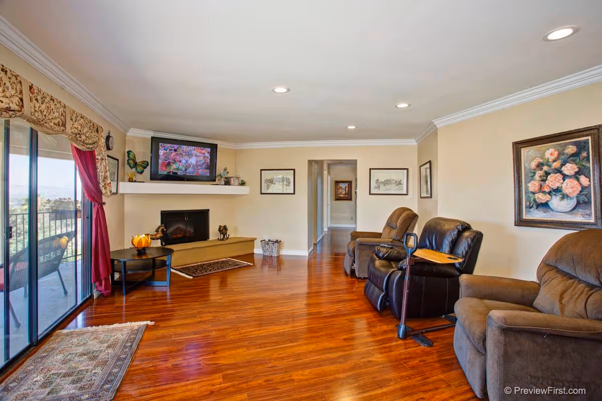 A spacious living room with polished wooden floors, beige walls, and recessed ceiling lights. The room features three brown recliner chairs, a wall-mounted TV above a fireplace, framed artwork on the walls, and a sliding glass door with floral curtains leading to an outdoor balcony with seating.