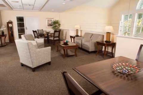 Cozy senior living common area with sofas, chairs, tables, and soft lighting.