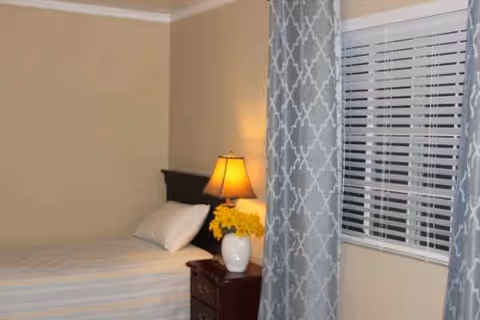 A bedroom with a bed, nightstand, lamp, flowers, and patterned curtains by a window.