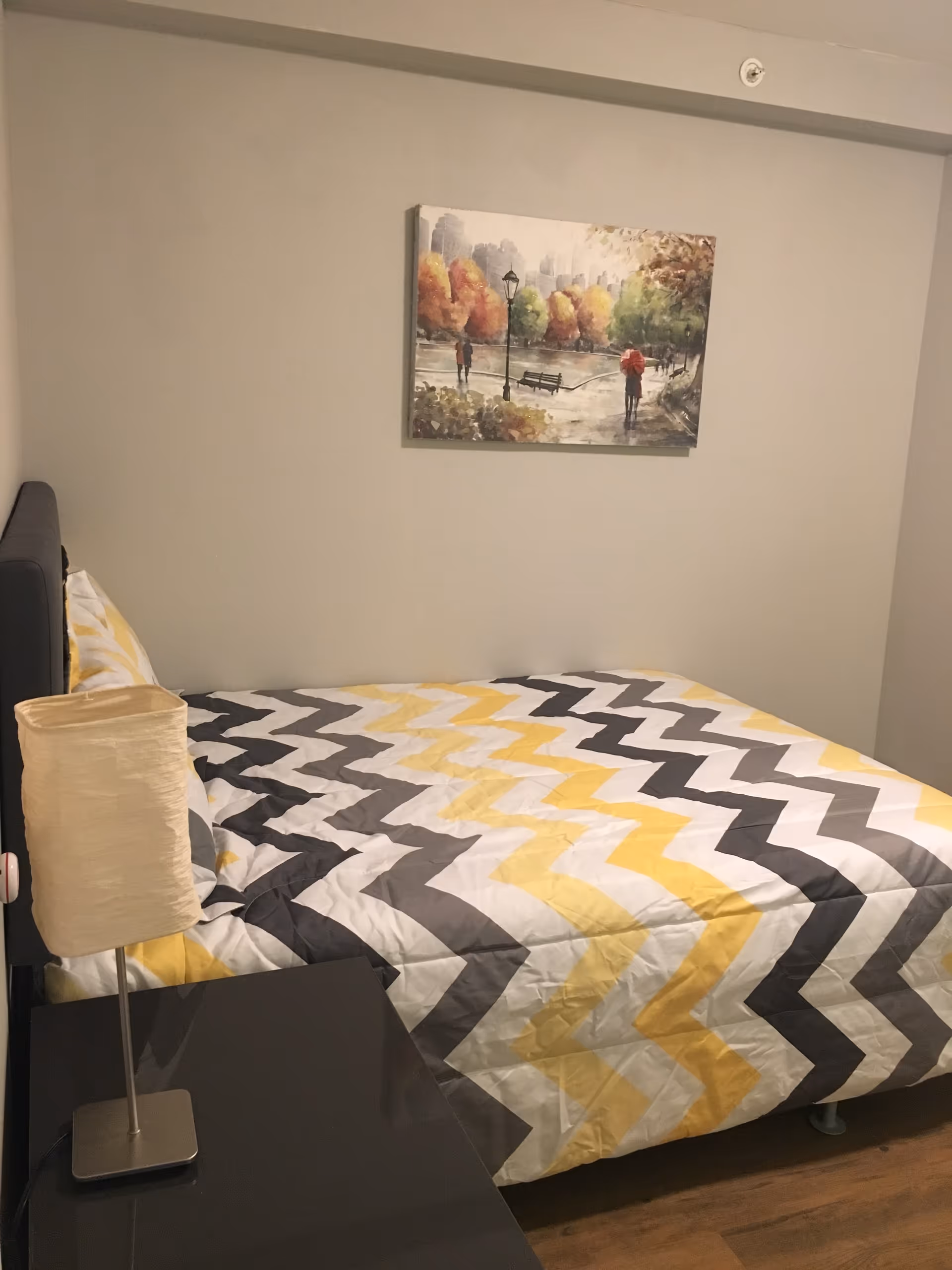 A bedroom with a bed covered in a zigzag patterned quilt in yellow, gray, and white. There is a small bedside table with a lamp on it next to the bed. A painting depicting a park scene with people walking and autumn trees hangs on the wall above the bed.