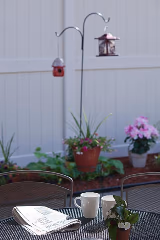 Patio table with a newspaper, two mugs and a small potted plant in front of a fenced garden with hanging bird feeders and flowers.