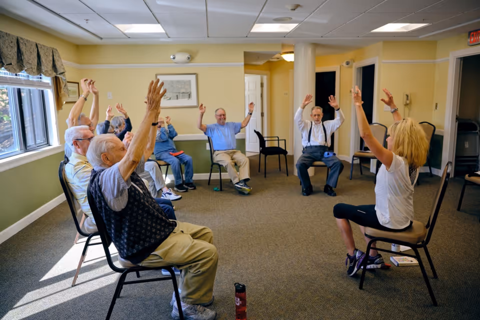 A group of elderly people seated in a circle in a room, participating in a seated exercise session led by a woman. They all have their arms raised above their heads. The room has beige walls, carpeted floor, and windows with curtains letting in natural light.