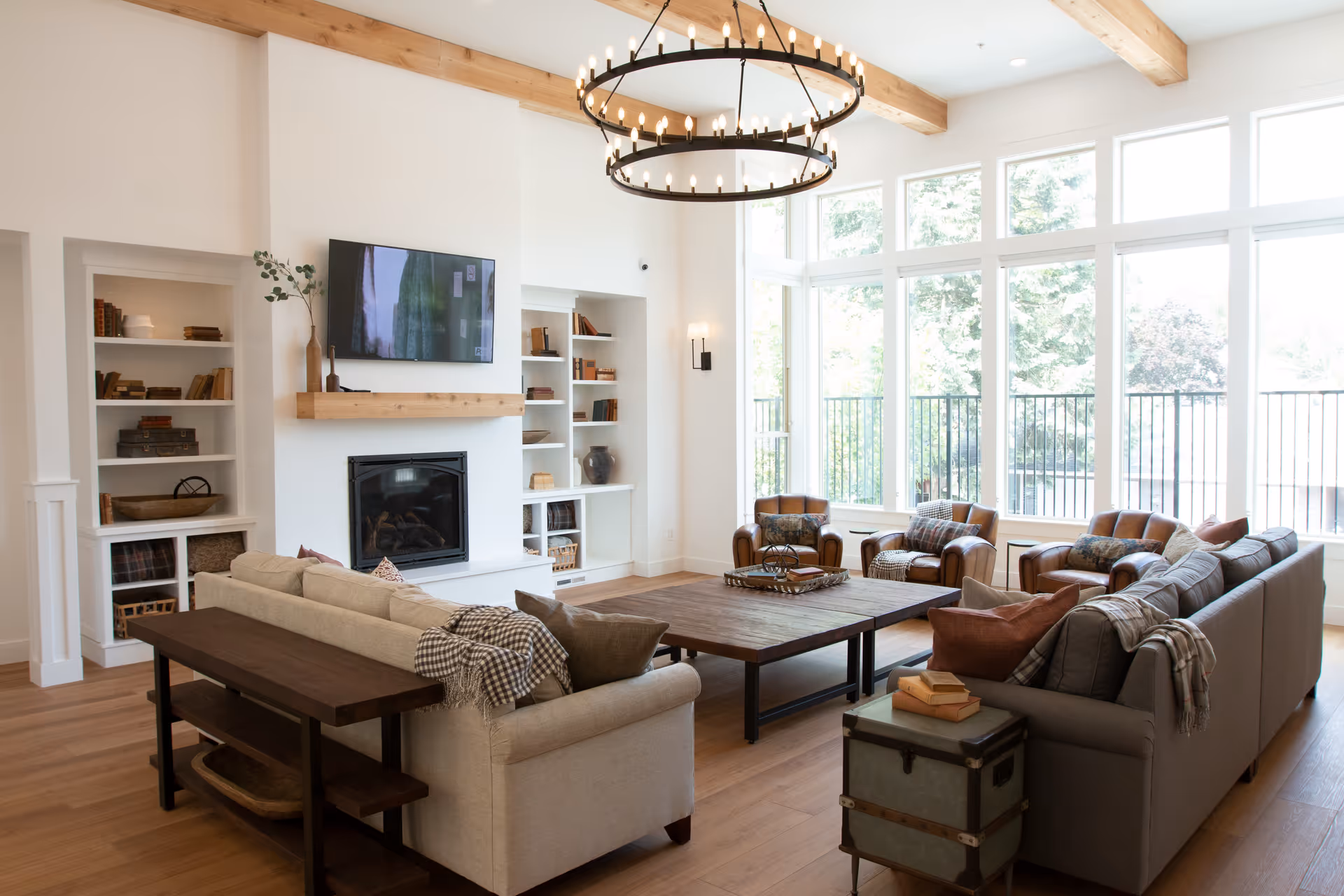 A bright and spacious living room with large windows letting in natural light. The room features two large sofas, three armchairs, a wooden coffee table, built-in white shelves with books and decorative items, a wall-mounted TV above a fireplace, and a large circular chandelier hanging from a ceiling with wooden beams.
