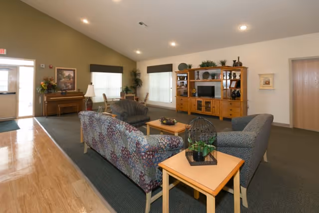 A spacious living room area with patterned sofas and armchairs arranged around a wooden coffee table and side table. The side table has a decorative birdcage with a plant inside. In the background, there is a large wooden entertainment center with a TV and various decorative items. The room has carpeted flooring, large windows with blinds, and a piano near the entrance door.
