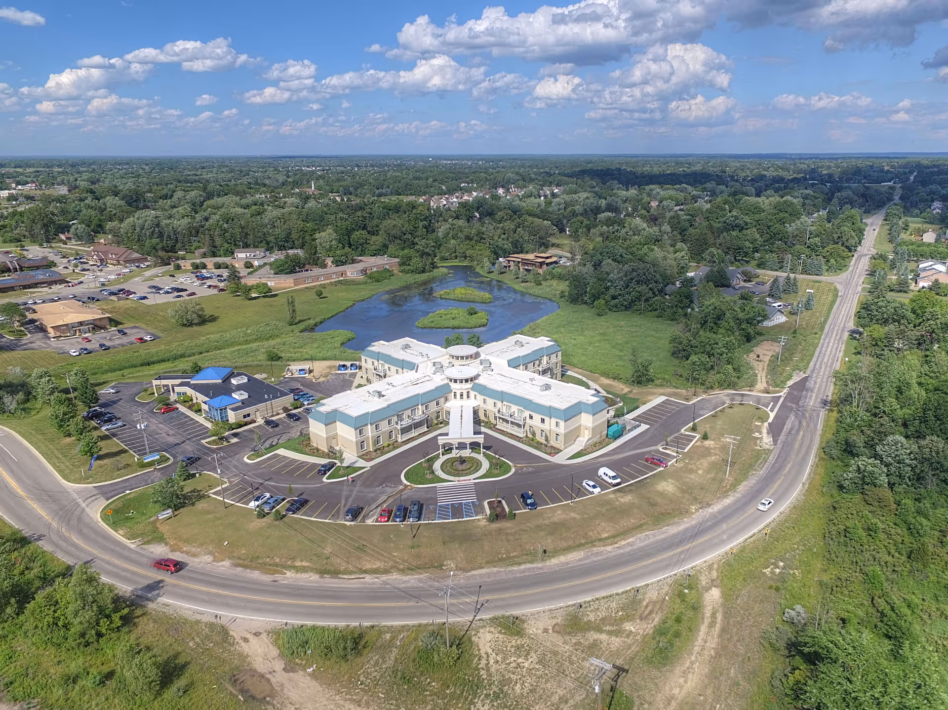 Aerial view of Tranquility Estates, a senior living facility with a large, cross-shaped main building surrounded by parking lots and greenery. The facility is situated near a pond and is bordered by roads and trees under a partly cloudy sky.