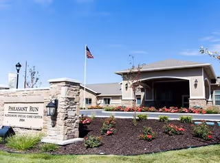 Front exterior of a single-story senior living facility with a stone entrance sign, landscaped flower beds, and an American flag.