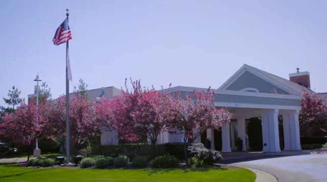 Exterior view of The Cortland Riverside Gardens facility with a white entrance structure supported by columns, surrounded by blooming pink trees and green landscaping. An American flag is flying on a flagpole in front of the building under a clear blue sky.
