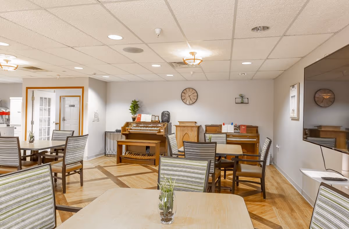 A well-lit common area with multiple tables and chairs arranged for seating. There are two pianos against the far wall, a clock above them, and a large flat-screen TV mounted on the right wall. The room has a light-colored ceiling with recessed lighting and wood-patterned flooring.
