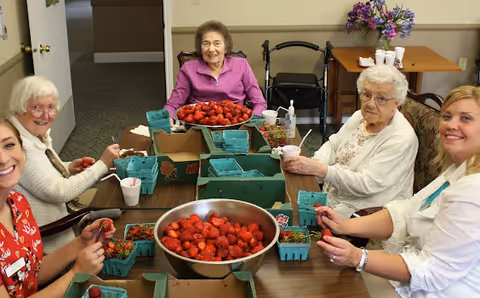 Five women sitting around a table indoors, smiling and holding strawberries. The table is filled with containers and a large bowl of strawberries. A walker and a small table with flowers and cups are visible in the background.