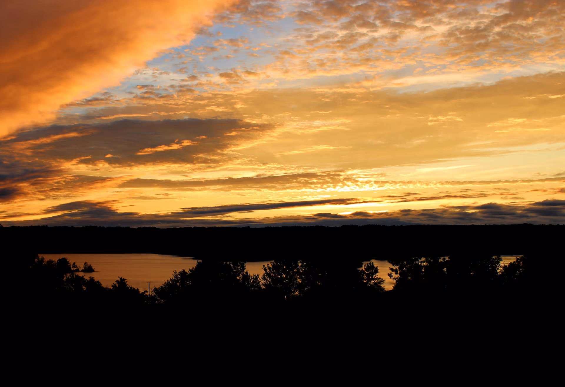 A scenic view of a lake at sunset with a vibrant sky filled with orange, yellow, and blue hues and scattered clouds above a dark silhouette of trees and land.