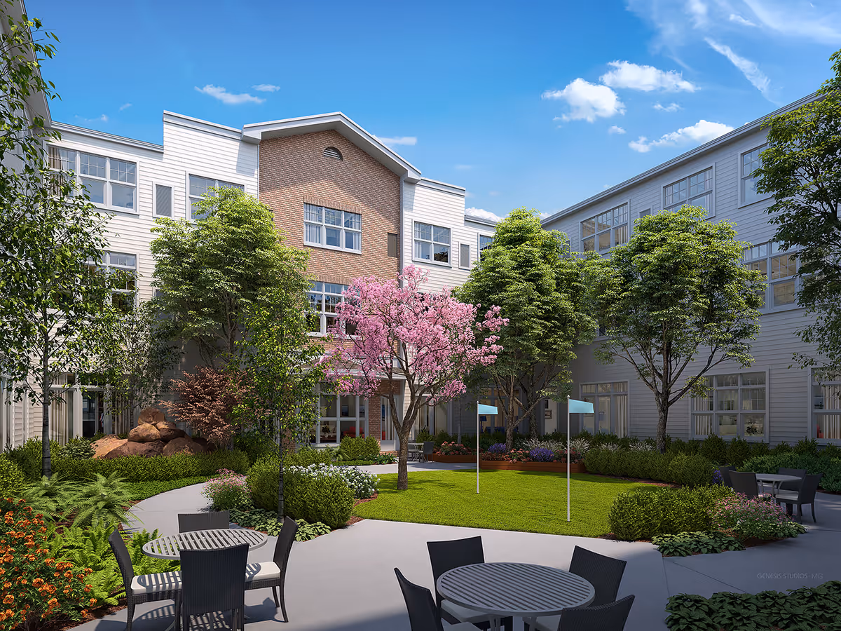 Outdoor courtyard area of The Sheridan at Eastside senior living facility featuring a well-maintained lawn, several trees including a blooming pink tree, shrubs, and flower beds. There are multiple round tables with chairs arranged on a paved patio area under a clear blue sky.