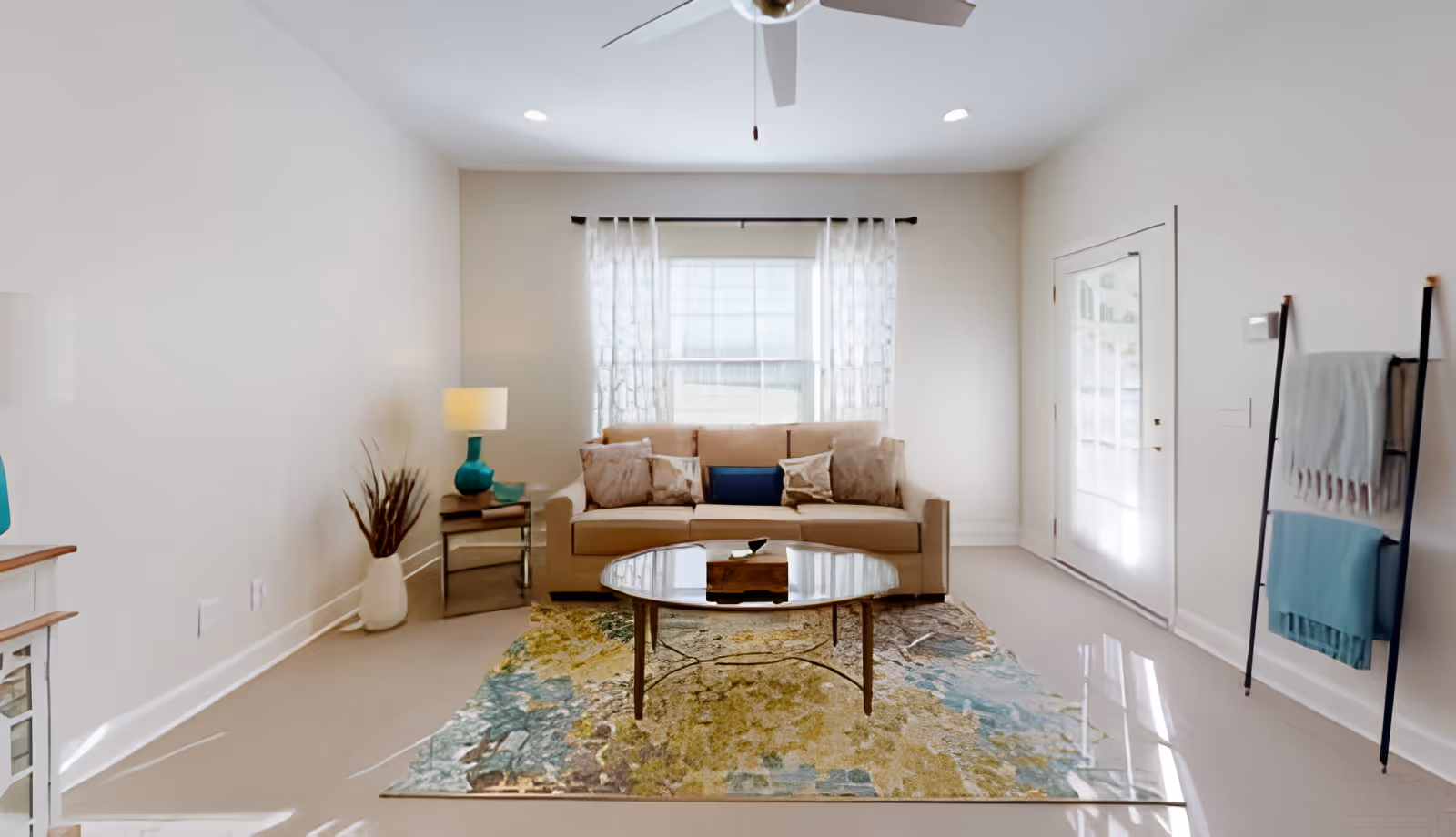 Bright living room with a sofa, coffee table on a patterned rug, side table lamp, and a blanket ladder near a glass door.