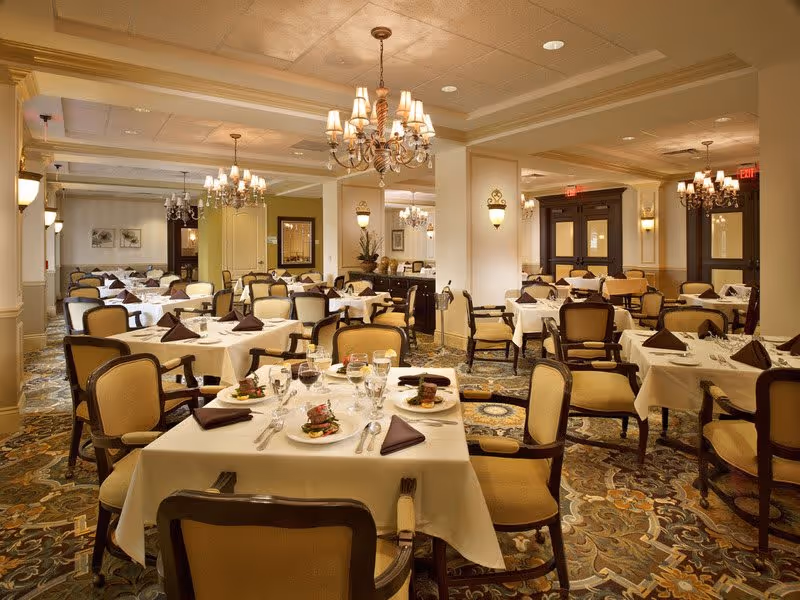 Elegant dining room with multiple tables covered in white tablecloths, each set with plates of food, glasses, silverware, and folded brown napkins. The room features upholstered chairs, ornate chandeliers, wall sconces, and a patterned carpet.