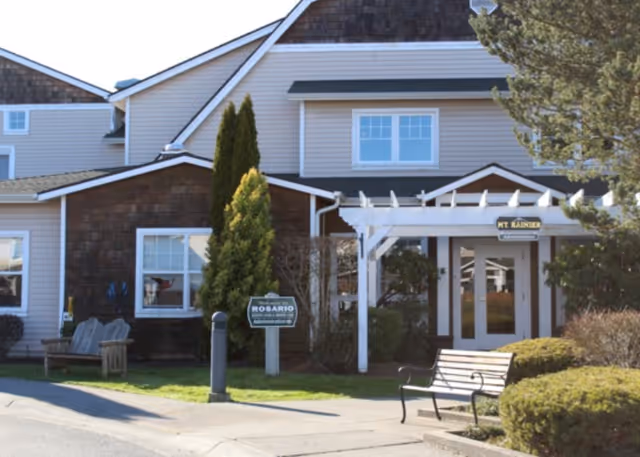 Front entrance of Rosario Assisted Living showing double doors under a white pergola, benches, landscaping, and a small sign.