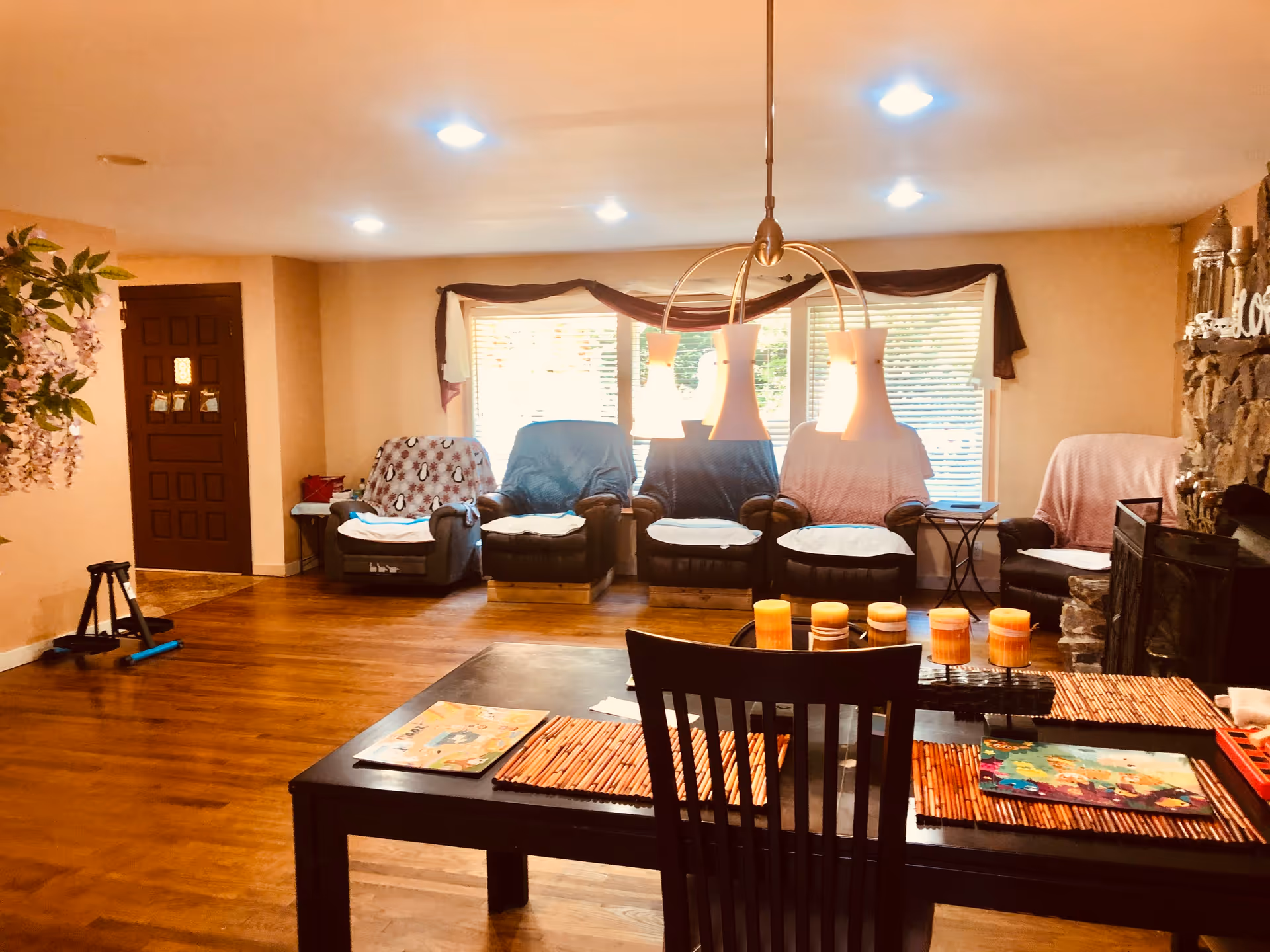 A cozy living room area with four recliner chairs covered with blankets arranged in front of large windows with blinds. A dark wooden table with placemats, candles, and magazines is in the foreground. The room has wooden flooring, warm lighting from ceiling fixtures, and a stone fireplace on the right side. A wooden door and a plant with purple flowers are visible on the left side.