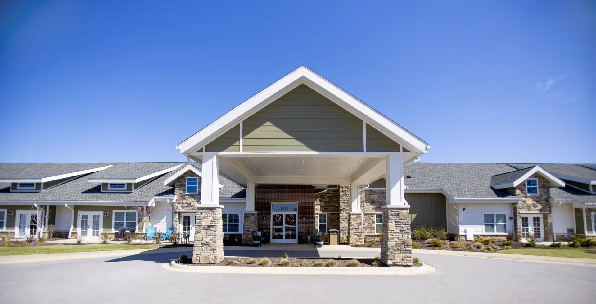 Front exterior view of a senior living facility named Ciel of Fayetteville with a covered entrance supported by stone pillars, a paved driveway, and a clear blue sky.