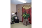 Small interior sitting area with a floor lamp, potted plants, a framed photo on a stand, and red drapes.