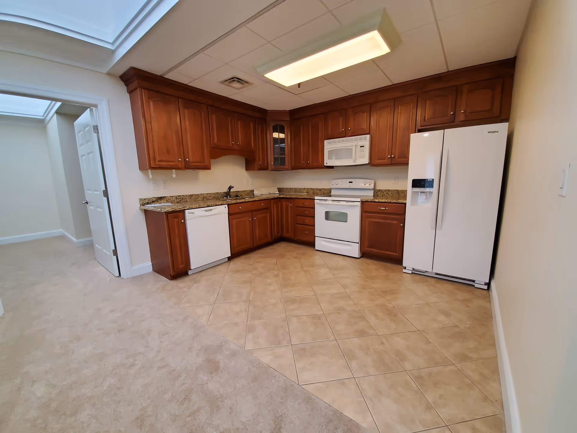 Interior view of a kitchen with wooden cabinets, granite countertops, white appliances including a refrigerator, stove, microwave, and dishwasher. The kitchen floor is tiled and opens into a carpeted area with a door leading to another room.