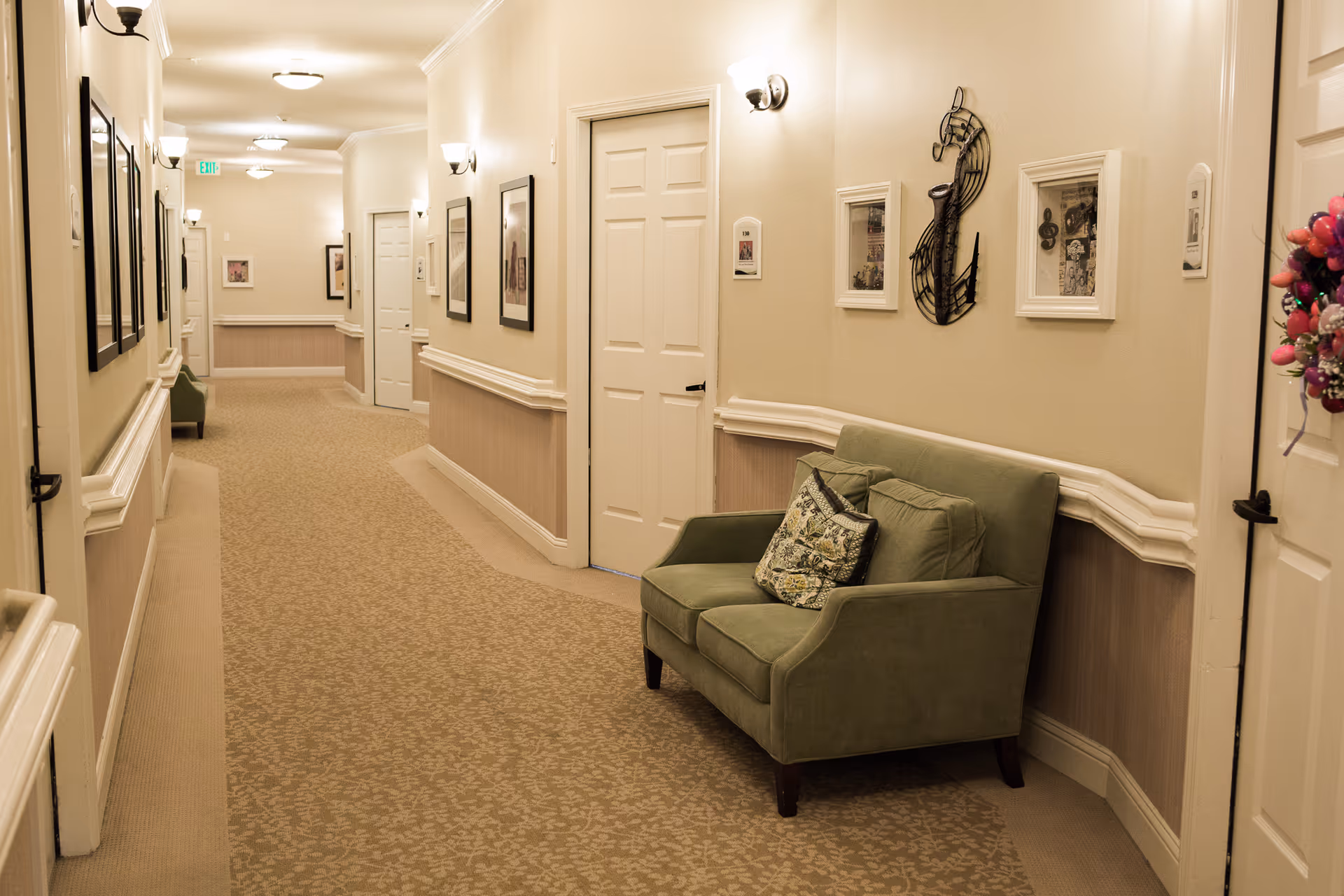 Carpeted hallway in a senior living facility with a green loveseat, framed artwork, wall sconces, and several doors.