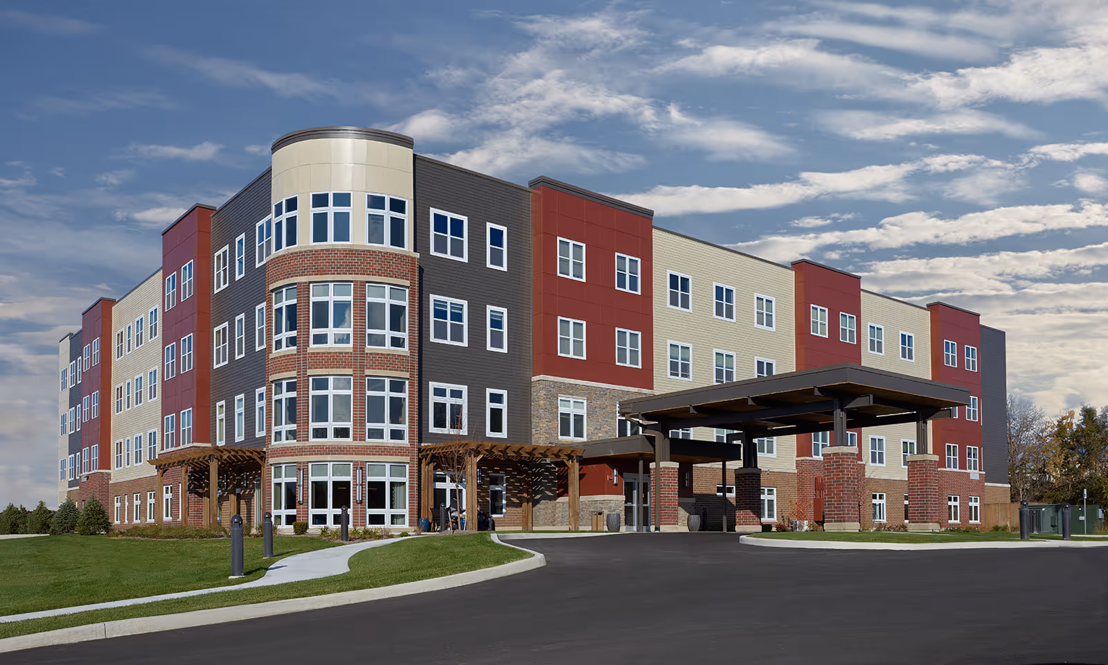 Four-story senior living building with a covered entrance, mixed brick and siding facade, and a landscaped curved driveway under a partly cloudy sky.