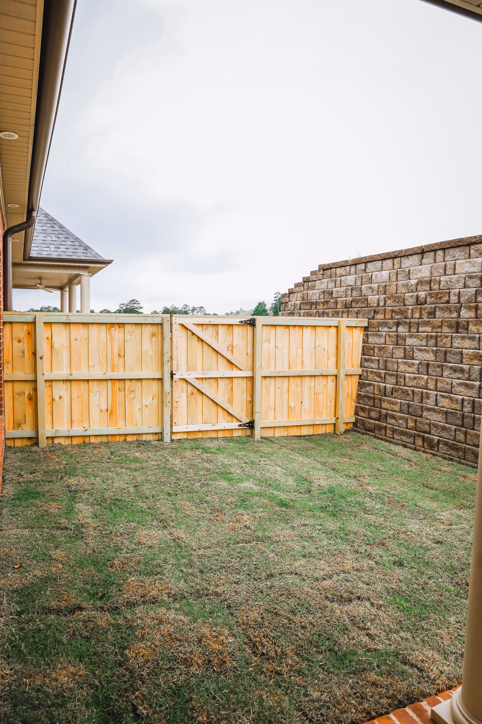 Small fenced backyard with a wooden gate, grassy lawn, and a stone retaining wall beside a house.
