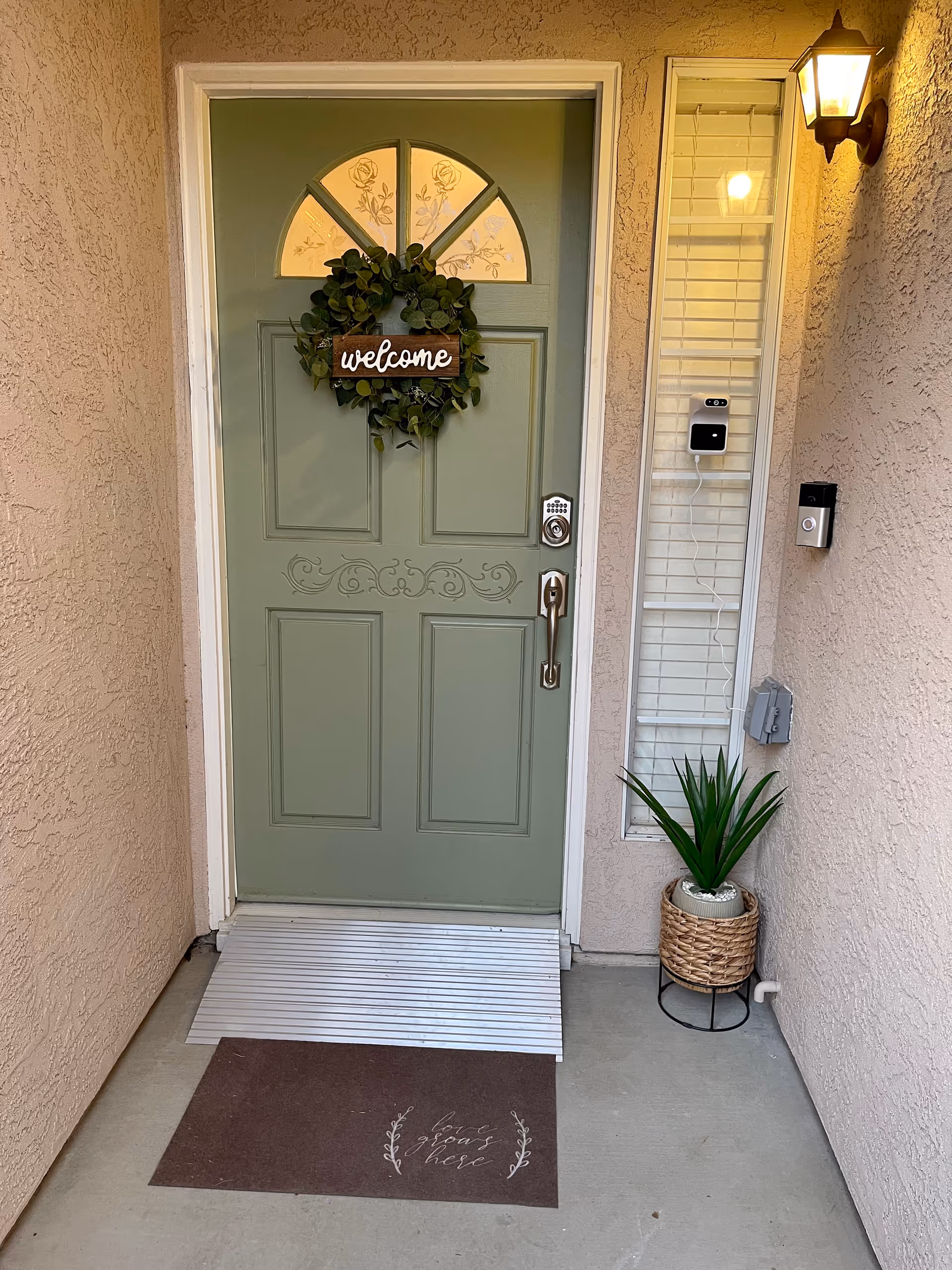 Front entrance of a home with a green door decorated with a wreath and a wooden sign that says 'welcome'. There is a brown doormat with the words 'love grows here', a small potted plant in a woven basket on a stand, a doorbell, a keypad lock, and a wall-mounted light fixture illuminating the area.