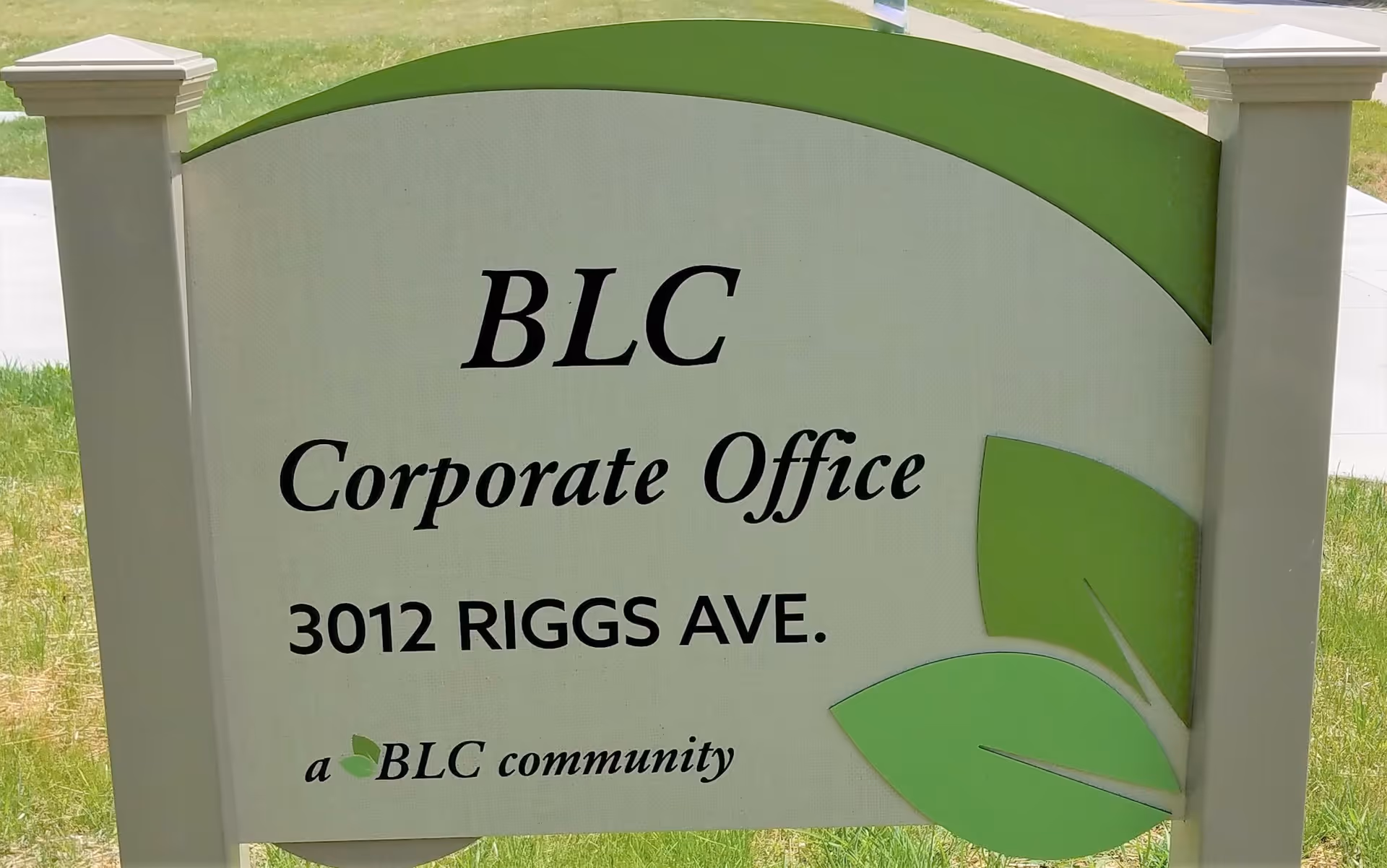 Outdoor sign for BLC Corporate Office at 3012 Riggs Ave, with green leaf graphics on the right side of the sign, standing on a grassy area with a sidewalk in the background.