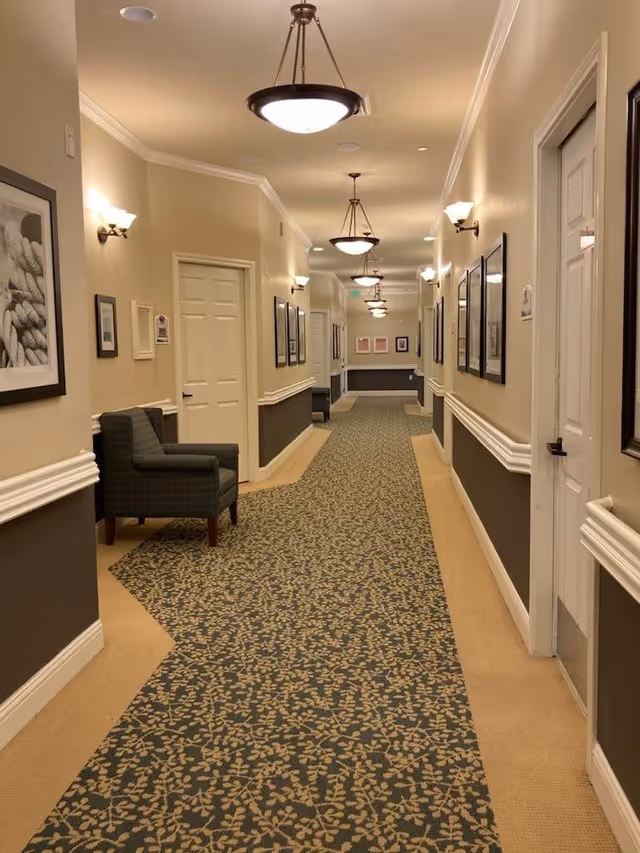 A long, well-lit hallway in a senior living facility with patterned carpet and beige walls. The hallway is decorated with framed pictures and has several closed white doors on both sides. There are wall-mounted lights and ceiling light fixtures evenly spaced along the corridor. A single upholstered chair is placed against the left wall.