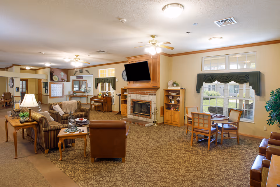 A spacious living room area in an assisted living facility featuring a fireplace with a mounted TV above it, several armchairs and sofas arranged around coffee tables, a piano against the wall, and a small round table with four chairs near a window with green valances. The room is well-lit with ceiling lights and a ceiling fan, and decorated with plants and framed pictures.