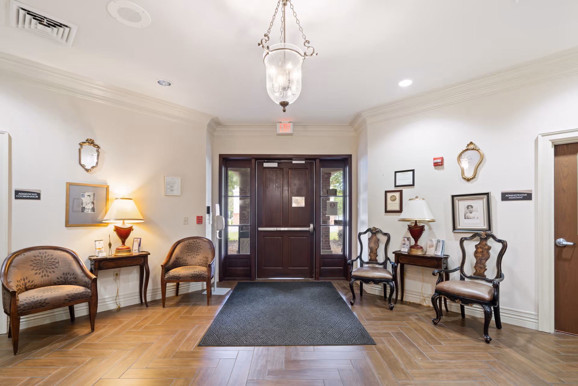 Entrance area of Forest Manor Nursing and Rehabilitation Center with a dark wooden double door, four chairs arranged along the walls, two small tables with lamps, framed pictures and certificates on the walls, and a hanging light fixture from the ceiling.