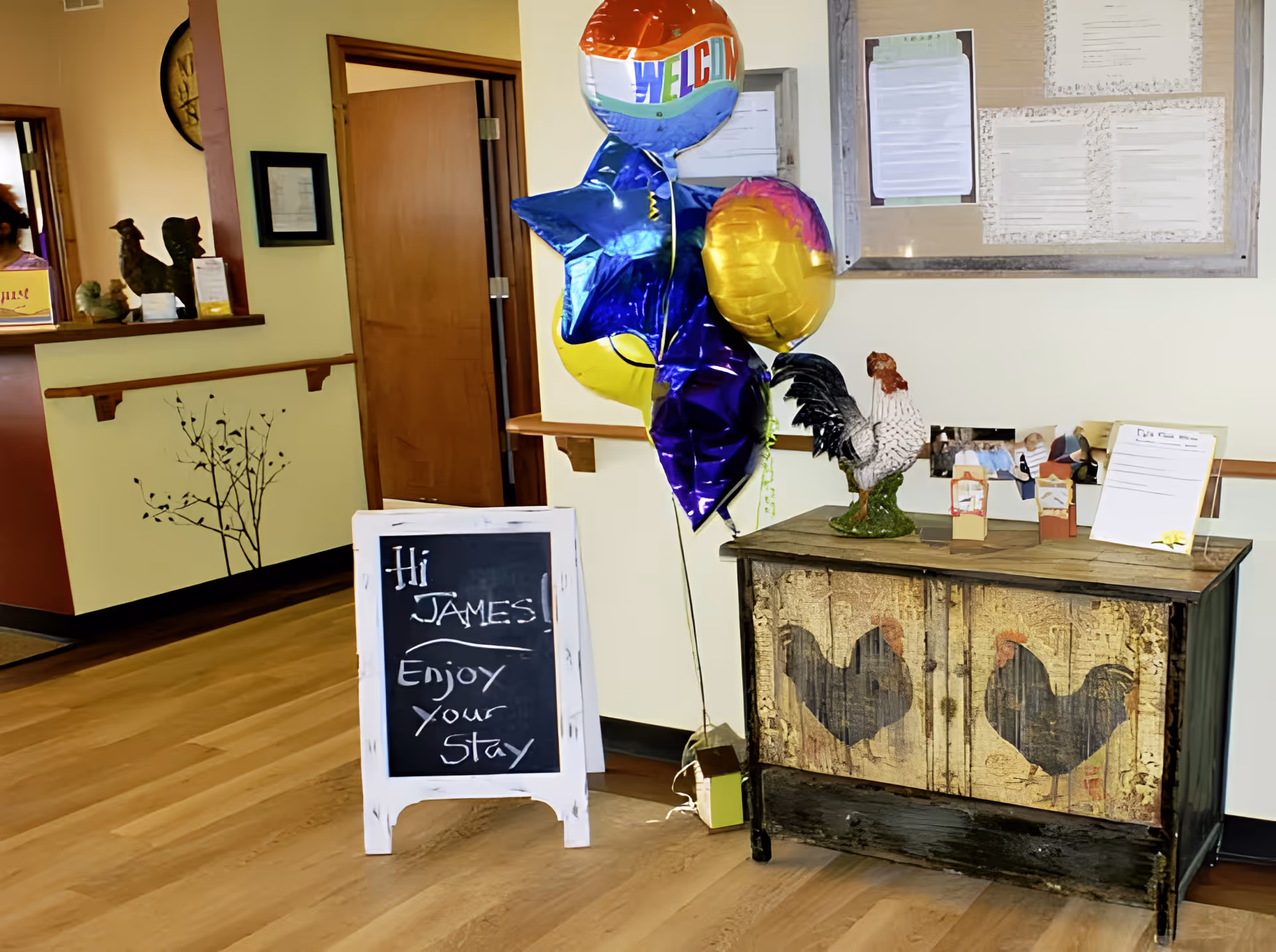 An interior hallway area with a wooden floor and light-colored walls. A small chalkboard sign reads 'Hi James! Enjoy your stay.' Next to the sign are colorful balloons and a rustic cabinet decorated with rooster images. A bulletin board with papers is mounted on the wall above the cabinet.