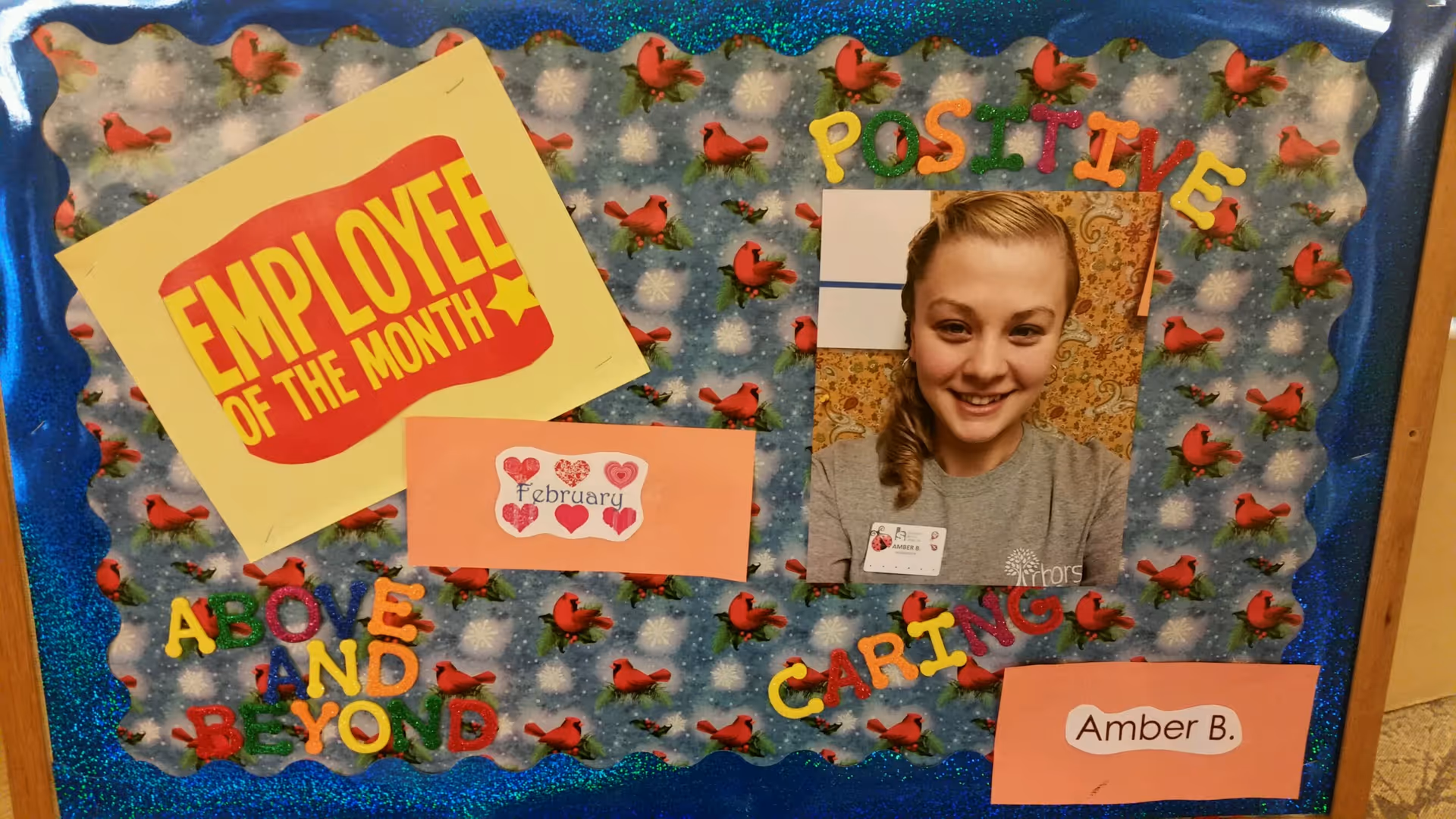 A decorated bulletin board with a photo of a smiling woman wearing a gray shirt and a name badge. The board has colorful letters spelling out 'EMPLOYEE OF THE MONTH', 'POSITIVE', 'ABOVE AND BEYOND', and 'CARING'. There are also small paper cutouts with the words 'February' and 'Amber B.'. The background of the board features a pattern with red birds and snowflakes.
