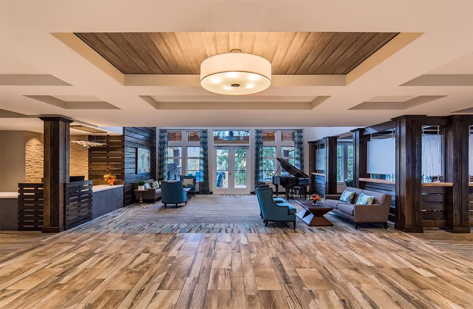 Spacious and well-lit common area in a senior living facility with wooden flooring, a large ceiling light fixture, comfortable seating including armchairs and sofas, a grand piano, and large windows with blue patterned curtains allowing natural light to enter.