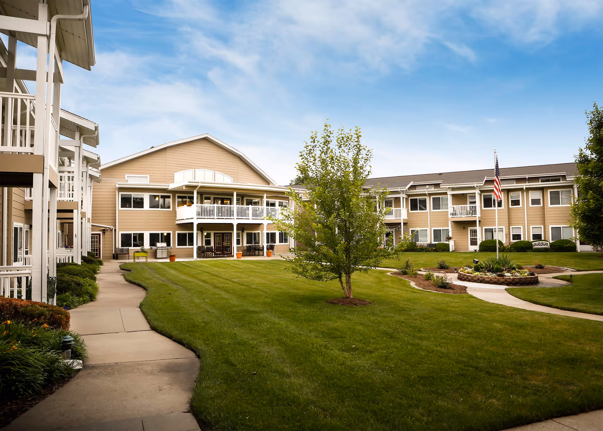 Exterior view of The Gardens at Barry Road Assisted Living and Memory Care facility showing a well-maintained lawn, a paved walkway, trees, and a two-story beige building with balconies and an American flag on a flagpole.
