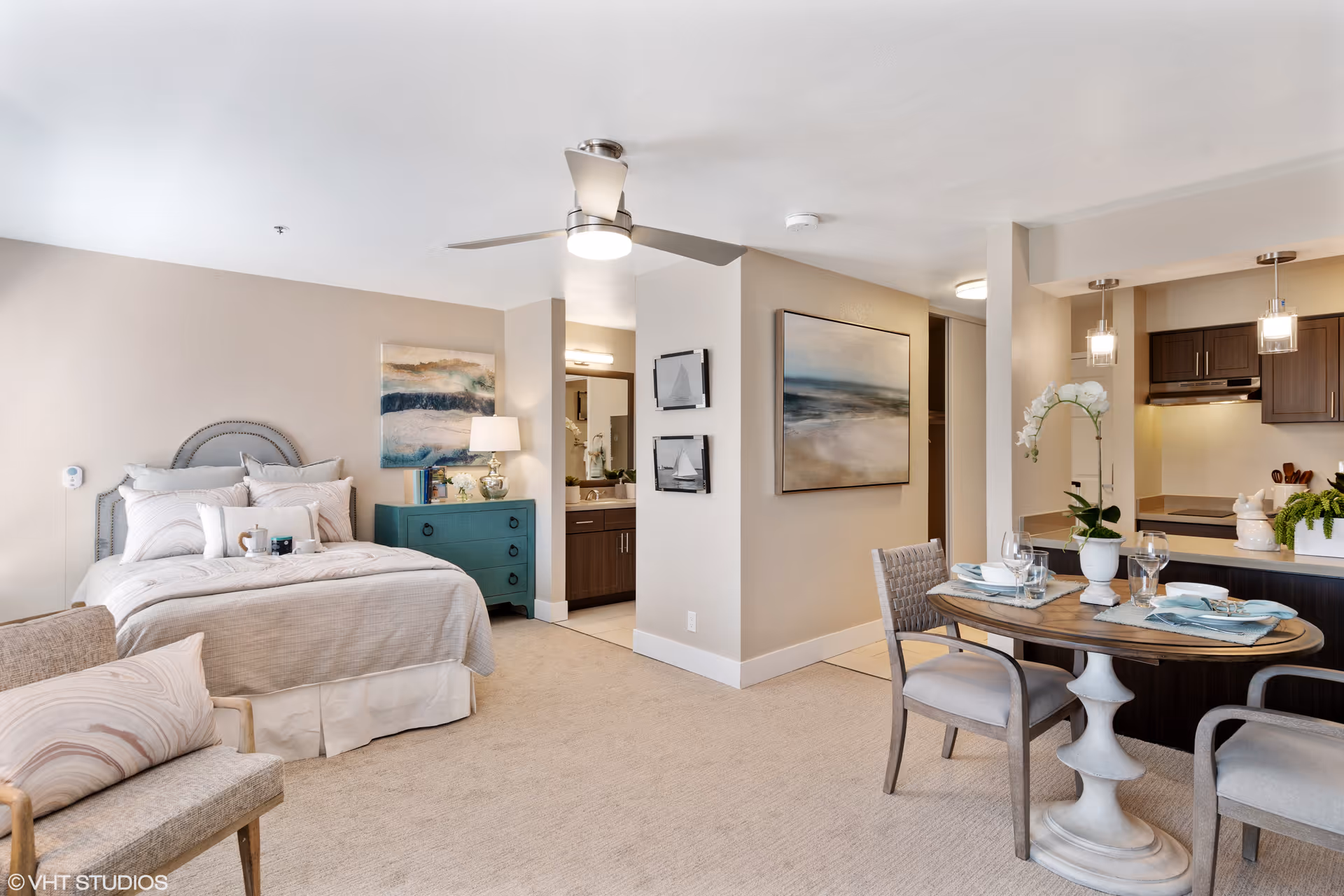 A bright and cozy studio apartment featuring a neatly made bed with multiple pillows, a teal chest of drawers with a lamp and decorative items, a small dining table set for two with plates and glasses, two chairs, and a modern kitchen area with dark cabinets and pendant lights. The room has beige walls, carpeted floor, ceiling fan, and several pieces of wall art.