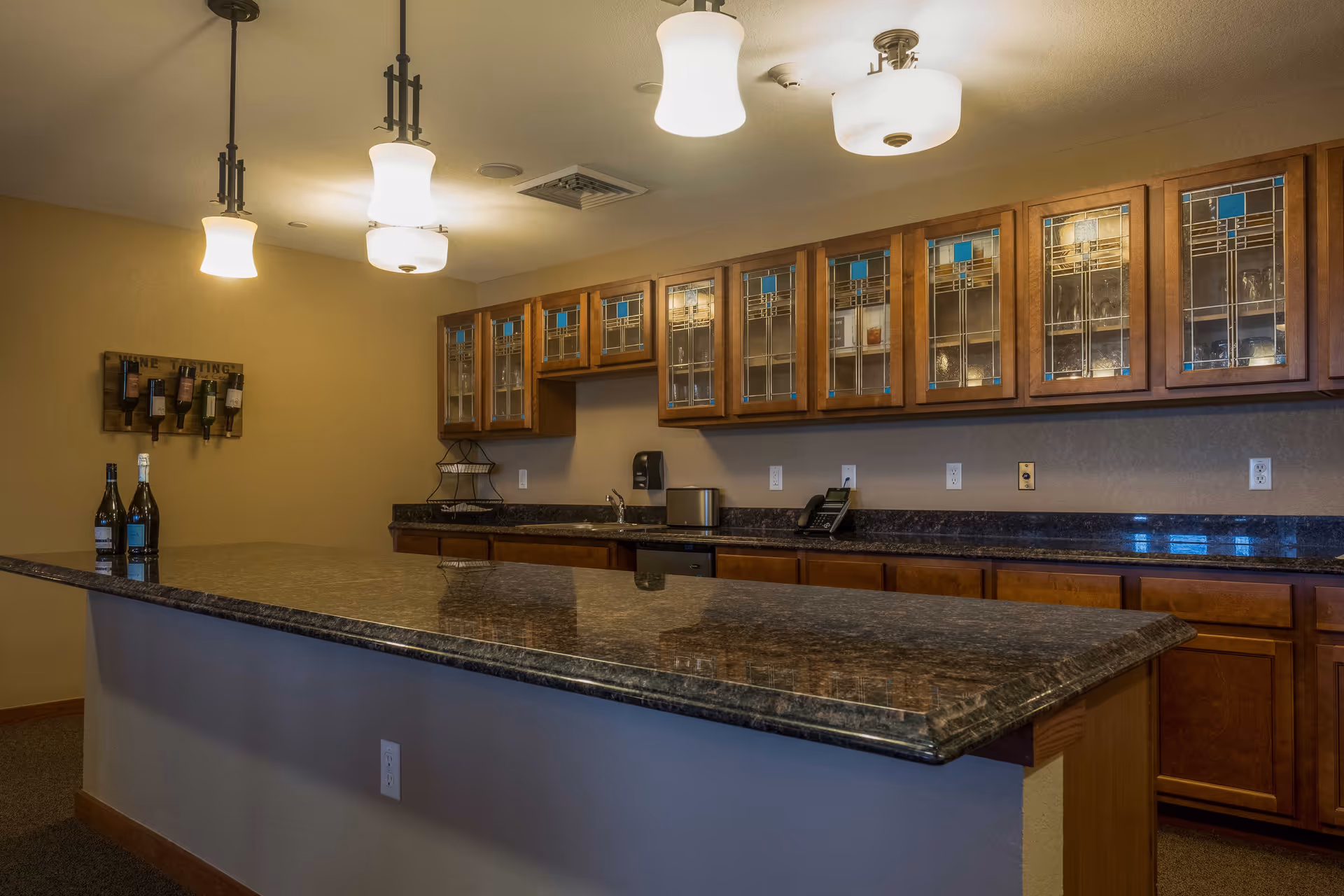 Interior view of a kitchen area with a long granite countertop island in the foreground, wooden cabinets with glass doors along the back wall, and three pendant lights hanging from the ceiling. There are bottles of wine on the countertop and a wine bottle holder mounted on the wall.