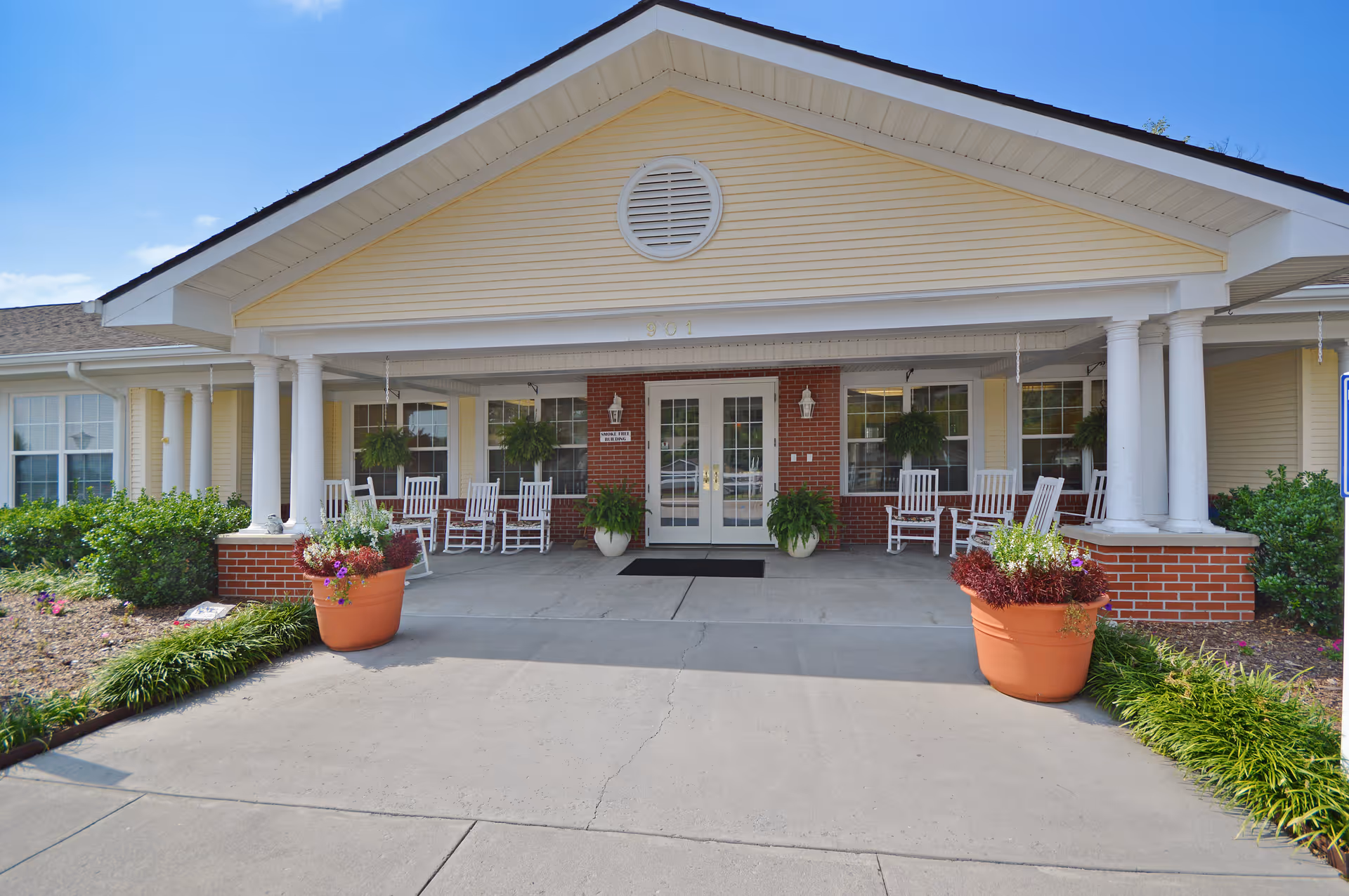 Front entrance of a senior living facility with a covered porch featuring white columns, several white rocking chairs, hanging plants, and large flower pots on either side of the walkway. The building has yellow siding with a brick base and double glass doors under a triangular pediment.