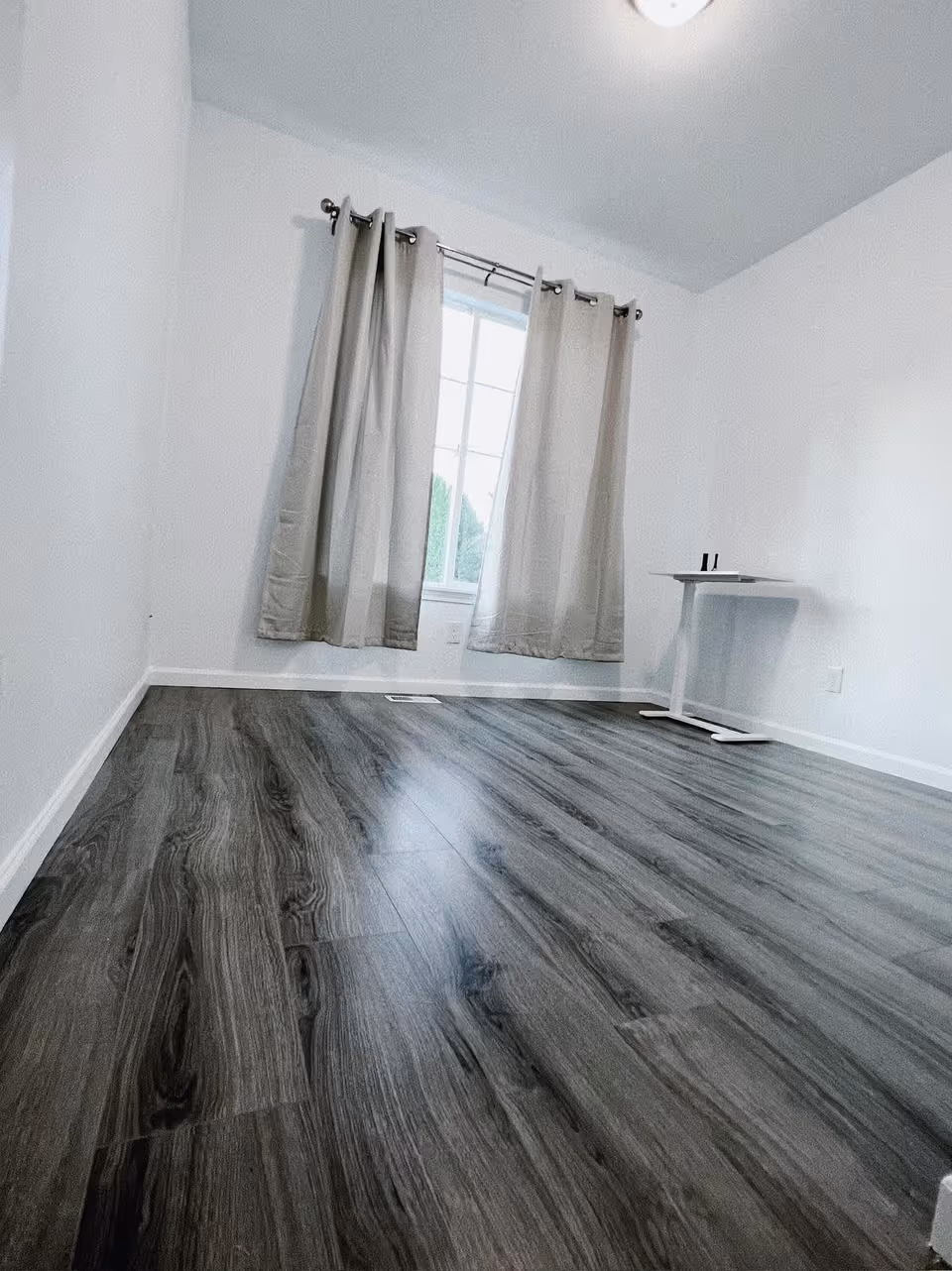 Empty room with gray wood flooring, white walls, a window with beige curtains, and a small white table in the corner.
