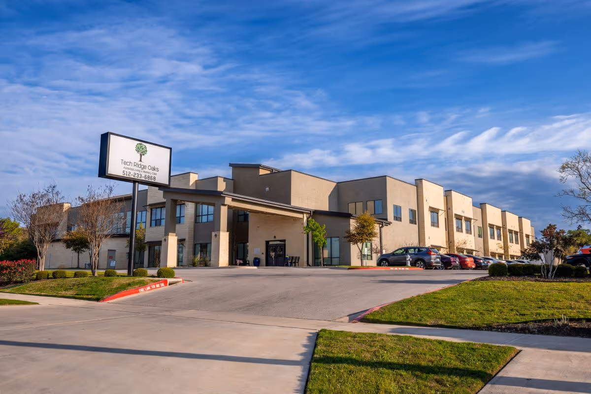 Exterior view of Tech Ridge Oaks, a two-story assisted living and memory care facility with a beige facade, large windows, a covered entrance, and a parking lot with several cars. The sky is partly cloudy with blue tones.