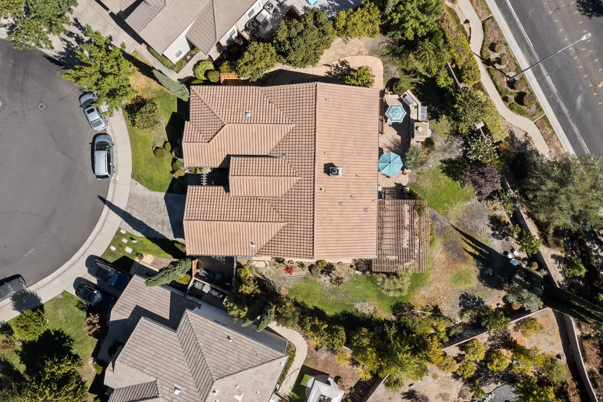 Aerial view of a residential facility with a large building featuring a tiled roof, surrounded by landscaped gardens, trees, pathways, and parked cars on a curved street.