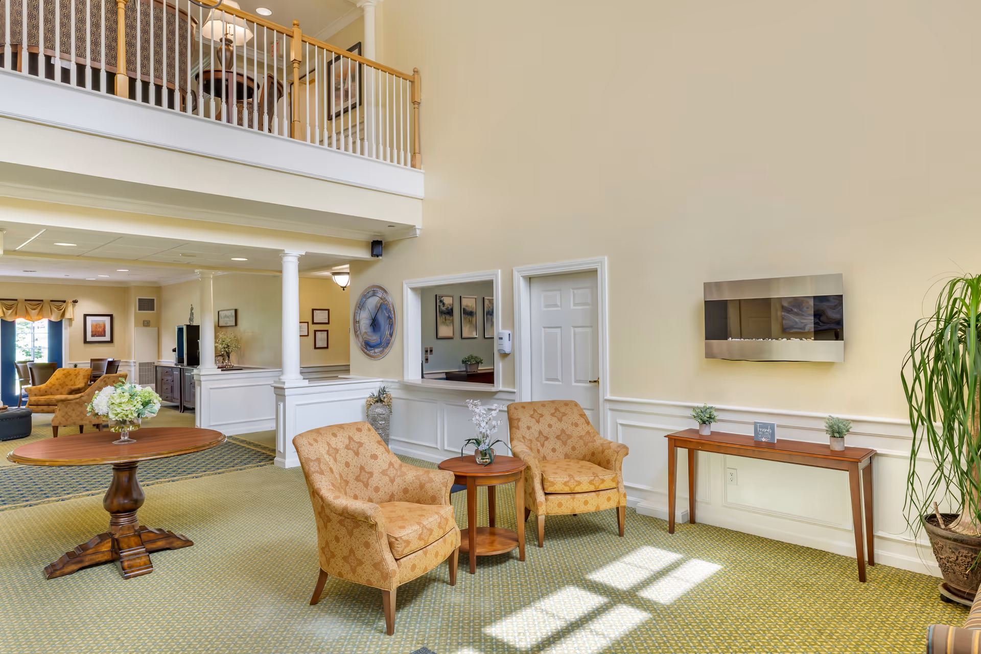 A bright and spacious common area in a senior living facility with two patterned armchairs and a small round wooden table between them. There is a wooden console table against the wall with small plants and a decorative sign. A modern wall-mounted electric fireplace is above the console table. The room has light yellow walls, green patterned carpet, and a high ceiling with a balcony railing visible above. In the background, there is more seating and framed artwork on the walls.