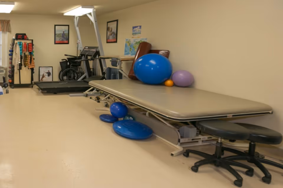 A physical therapy room with a large padded treatment table, various exercise balls in blue, purple, and orange, two black rolling stools, a treadmill, a wheelchair, and a rack holding exercise equipment such as weights and resistance bands. The walls are beige with framed pictures and a map.